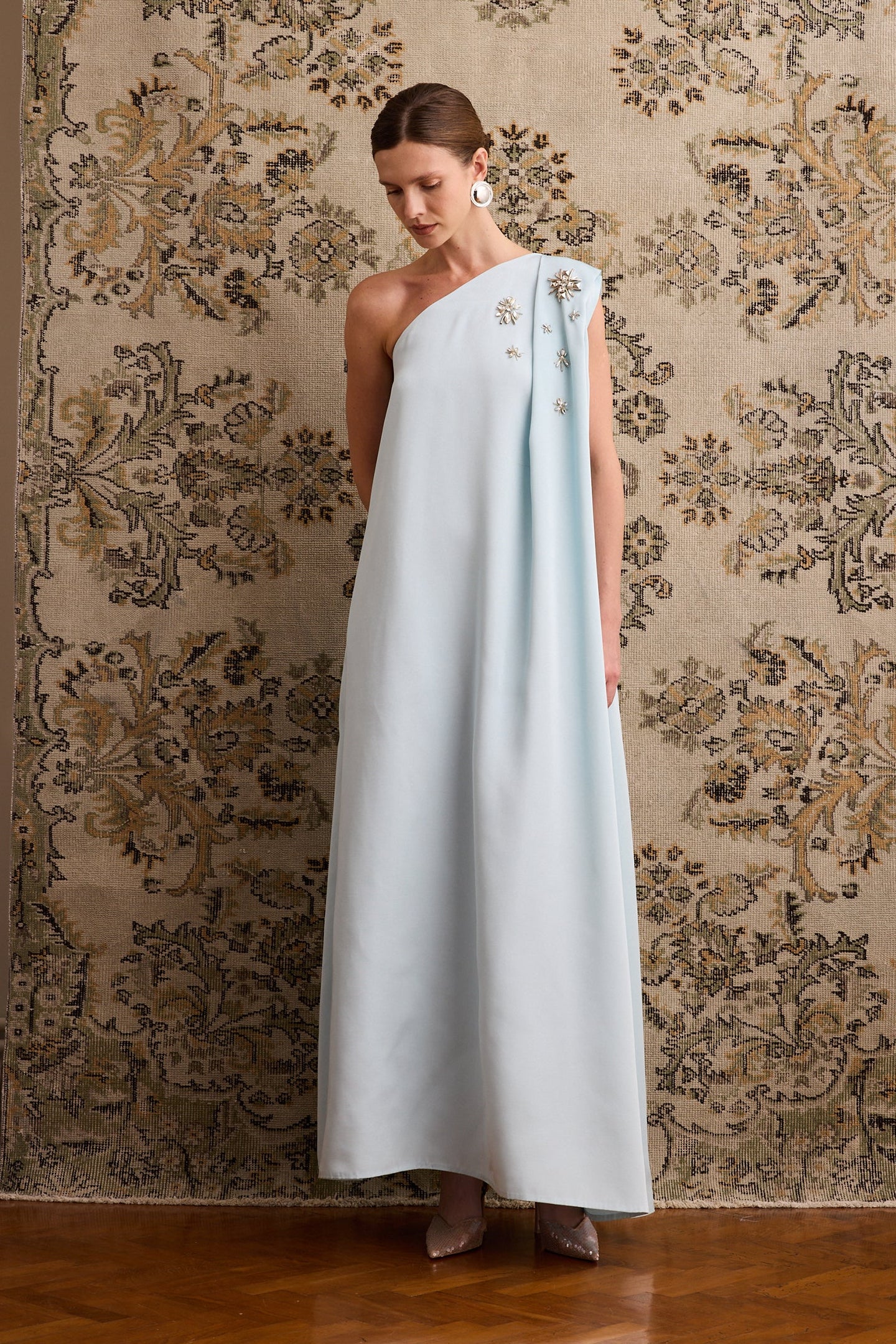 Woman wearing a light blue one-shoulder dress with floral embroidery against a patterned wall.