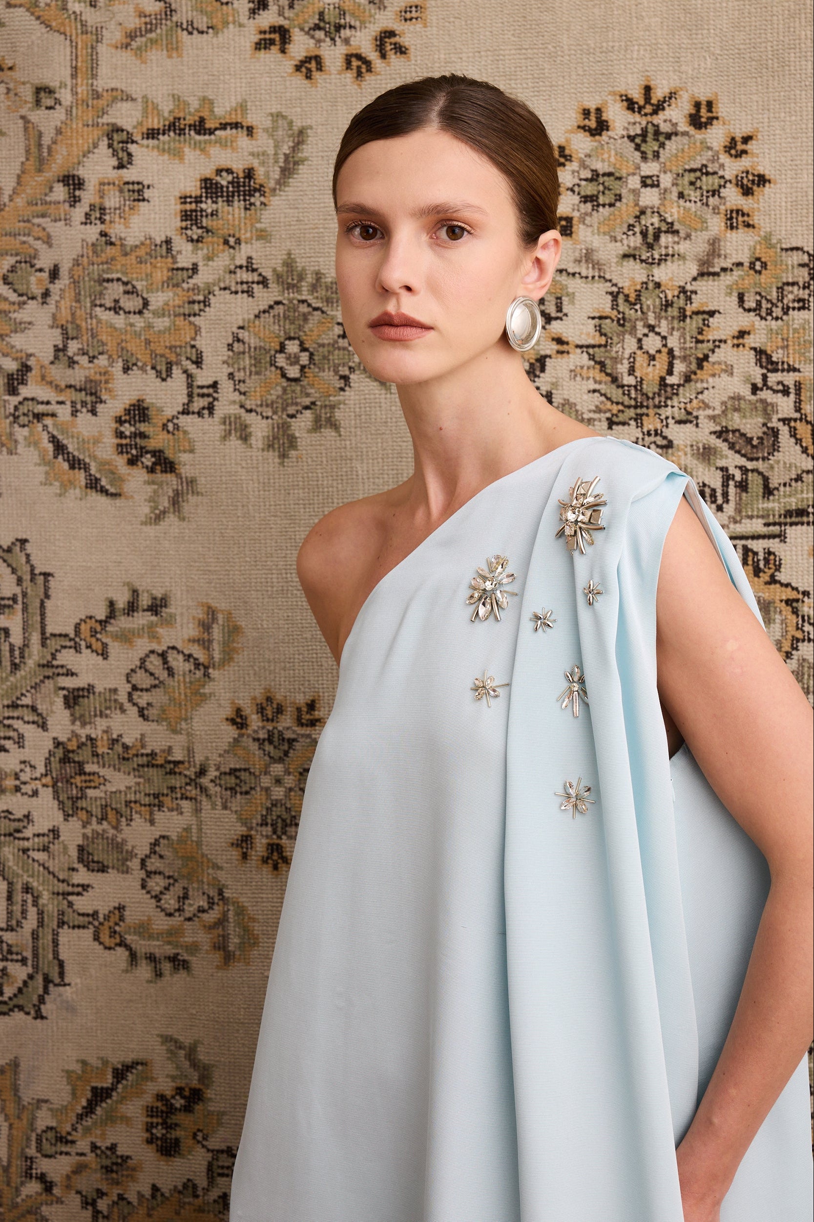 Woman wearing a light blue dress with floral patterns against a patterned wall.