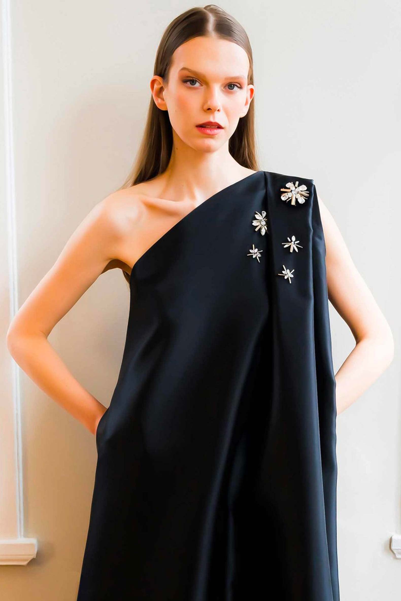 Woman wearing a black one-shoulder dress with floral embellishments against a neutral background